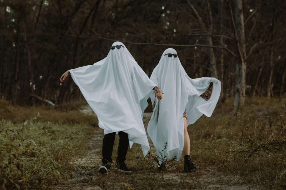 Couple wearing ghost costumes with sunglasses holding hands on a forest path — playful Halloween date-night photo.